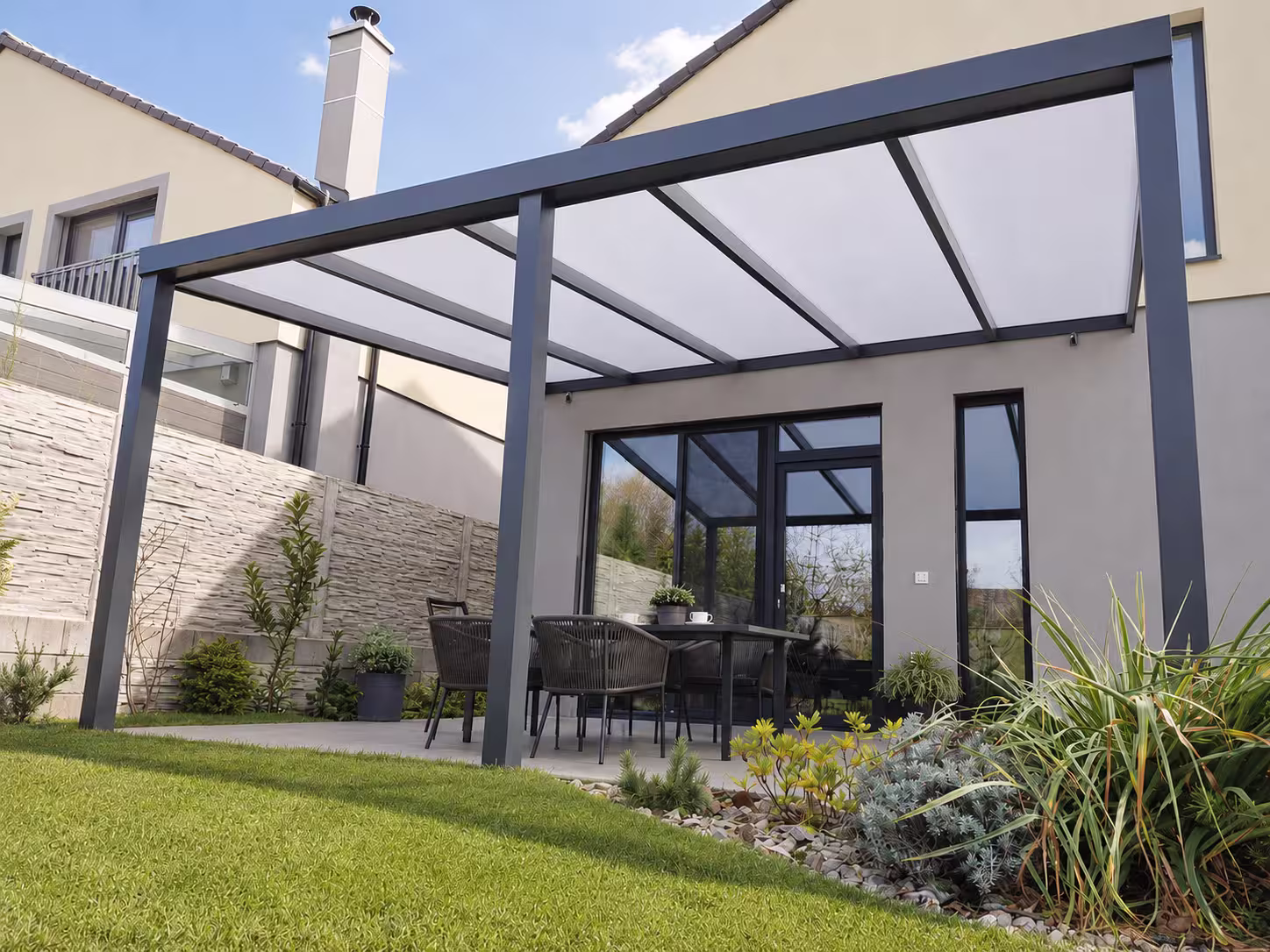 Anthrazitfarbene Aluminiumpergola Basic Class mit opalem Dach neben einem Haus mit Kamin