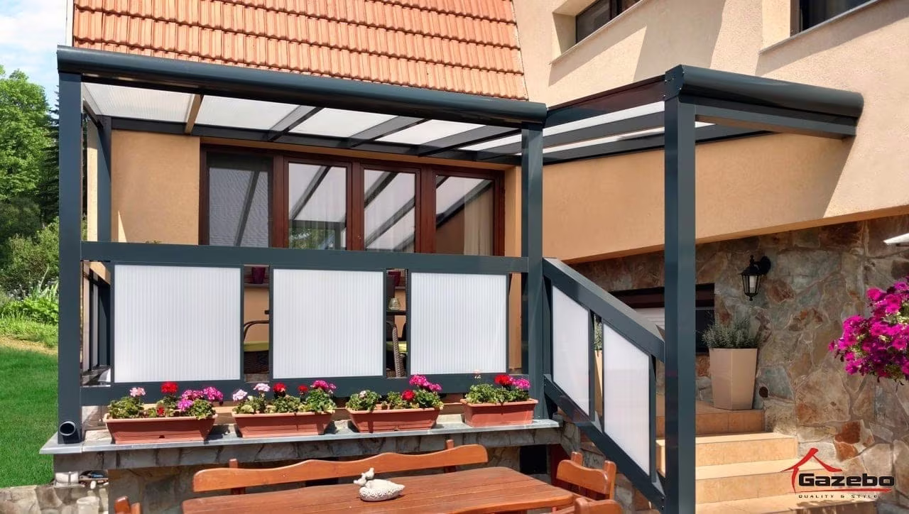 Anthrazitfarbene Aluminiumpergola Economic Class mit verglasten Seiten an einem Steinhaus mit Blumen