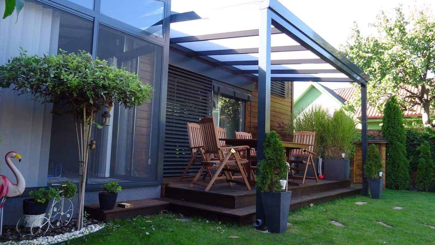 Anthrazitfarbene Aluminiumpergola Economic Class über einer Holzterrasse mit Essgruppe an einer Hecke