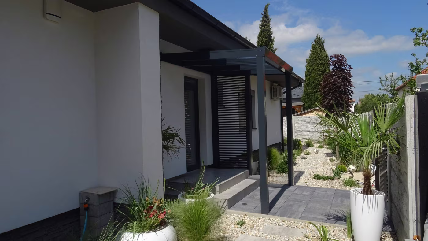 Anthrazitfarbene Aluminiumpergola Economic Class entlang eines weißen Hauses mit Topfpflanzen