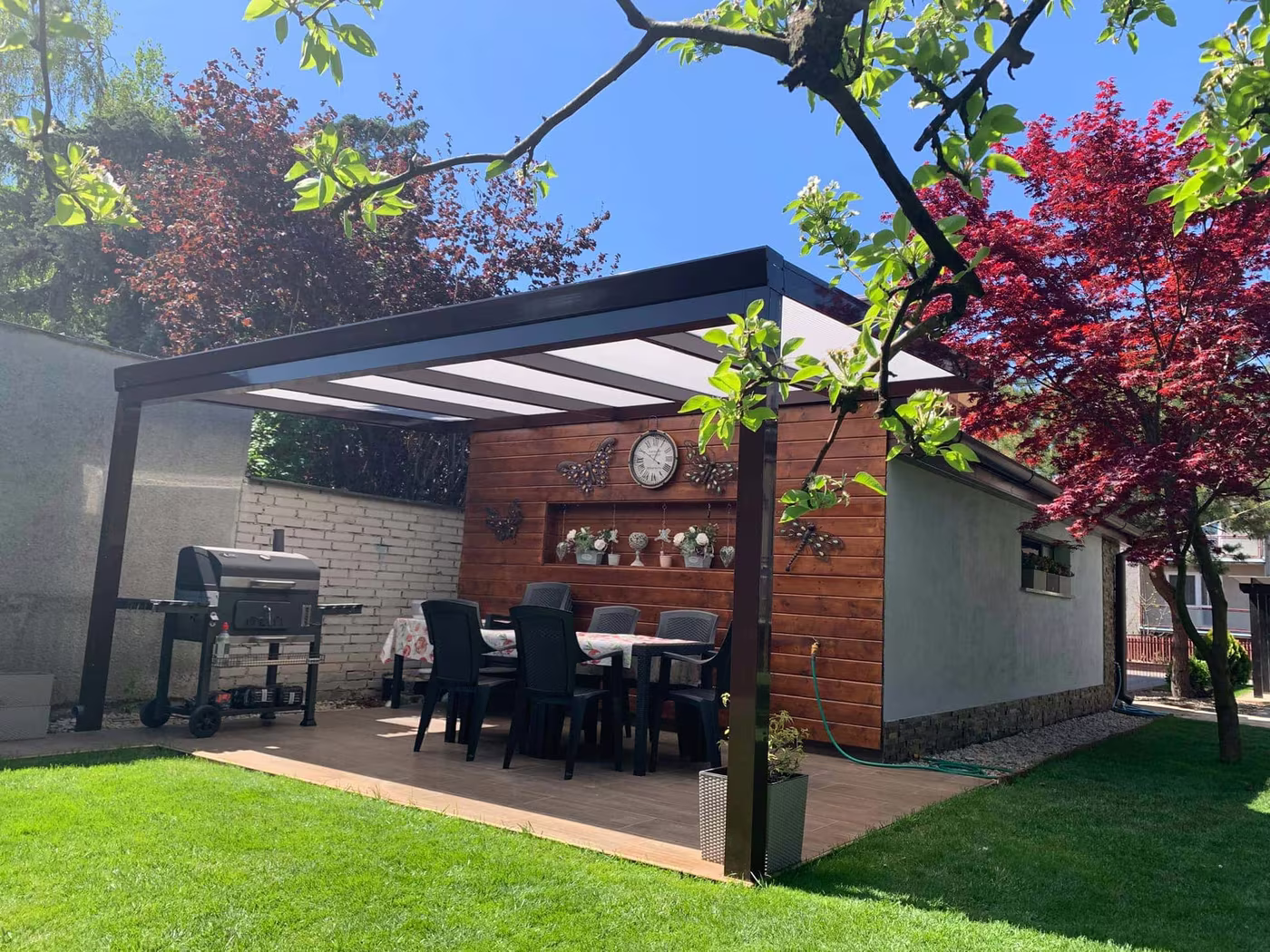 Anthrazitfarbene Aluminiumpergola Economic Class über einer gepflasterten Terrasse mit Grill und Sitzecke