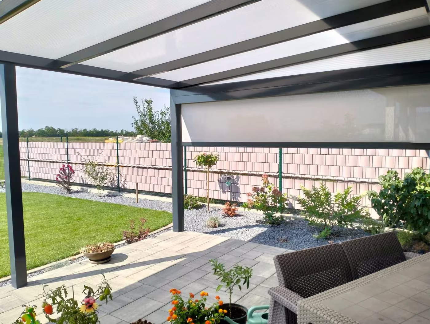 Anthrazitfarbene Aluminiumpergola Economic Class entlang eines Zauns mit Rattanmöbeln und Blumen