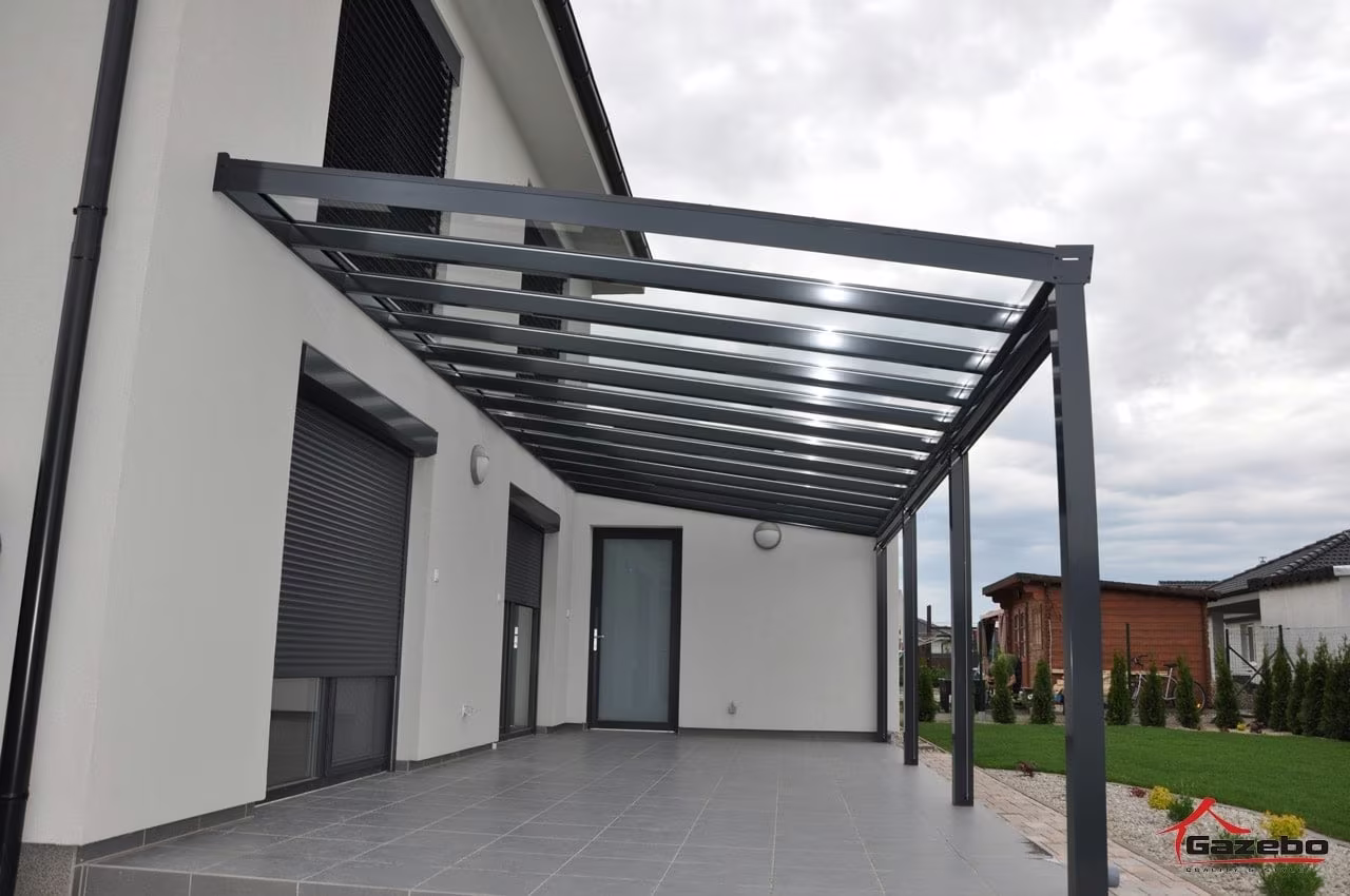 Anthrazitfarbene Aluminiumpergola Top Class über einer gepflasterten Terrasse entlang eines Hauses mit Rollläden