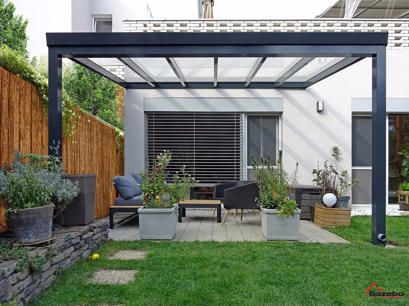 Anthrazitfarbene Aluminiumpergola Top Class über einer Terrasse mit Sitzgelegenheit an einem Haus mit Holzzaun