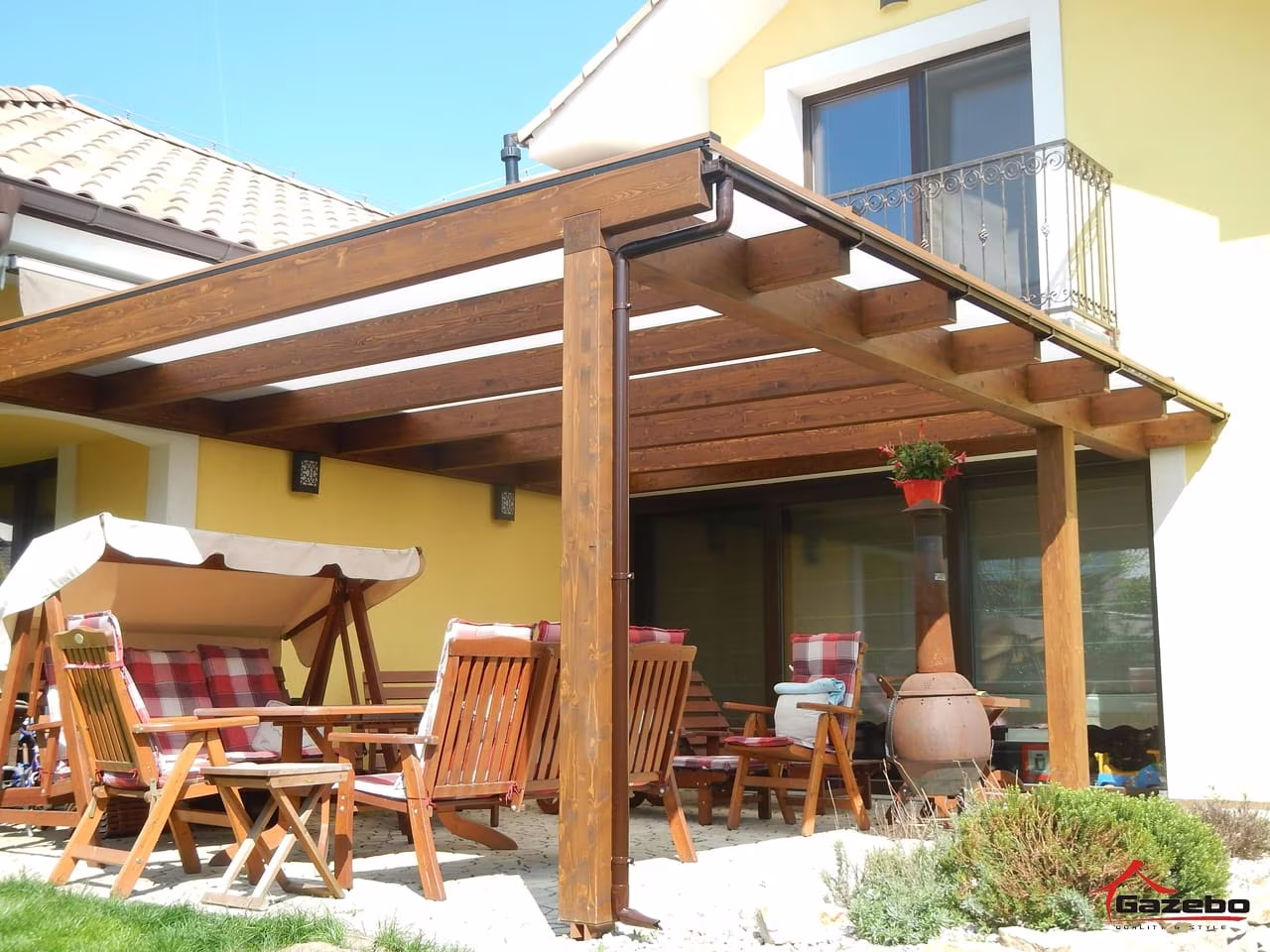 Holzpergola Fenix über einer Terrasse mit Gartenmöbeln und Hollywoodschaukel an einem gelben Haus