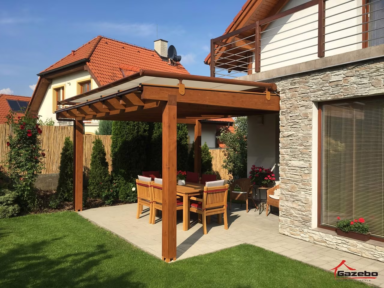Holzpergola Fenix über einer gepflasterten Terrasse mit Essgruppe an einem steinverkleideten Haus