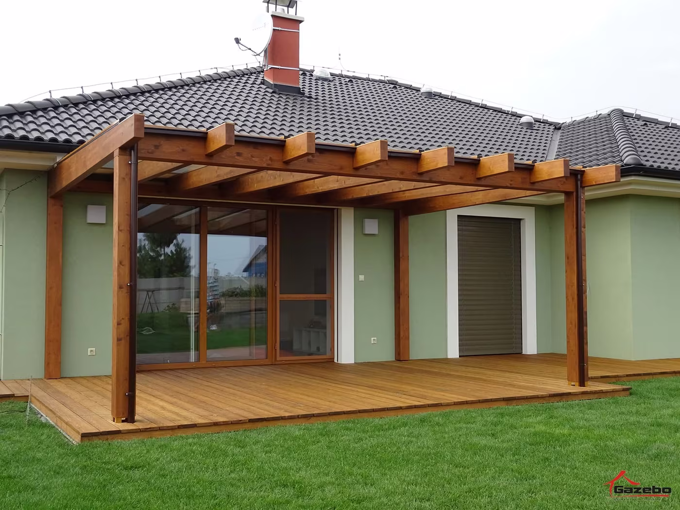 Holzpergola Fenix an einem Haus mit verglasten Türen über einer Holzterrasse mit Rasen